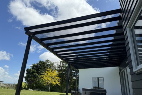 Black aluminium lean-to pergola with a clear roof attached to a dark weatherboard house