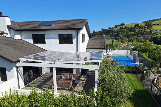 White pergola with a clear roof sheltering an outdoor lounge beside a two-storey home and pool